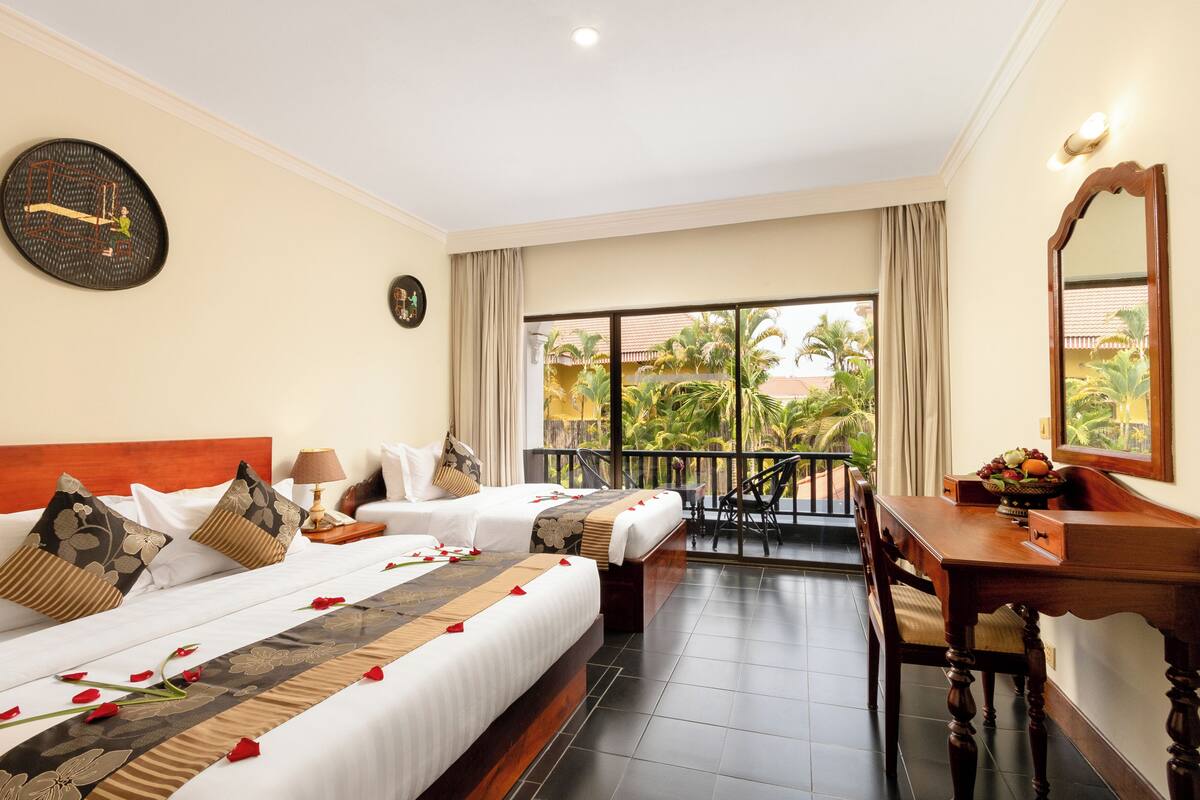 A hotel room with two beds, a balcony with a view of palm trees, a dining table, and a desk with a mirror.