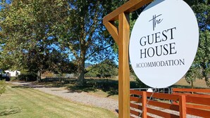 Schild für "The Guest House Accommodation" mit Holzzaun und Bäumen im Hintergrund.