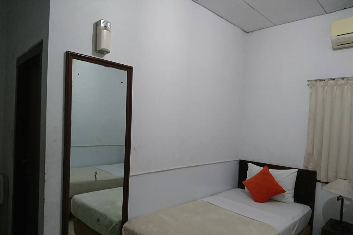 A small, simple room with a bed, a bedside table, a lamp, a mirror, and a wall-mounted air conditioner.