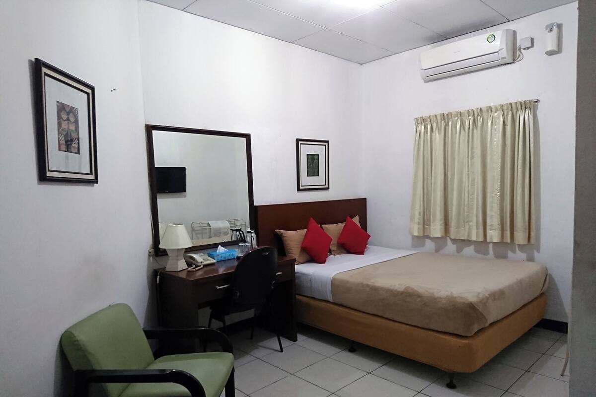 A hotel room with a bed, a desk with a chair, a mirror, and an air conditioning unit.