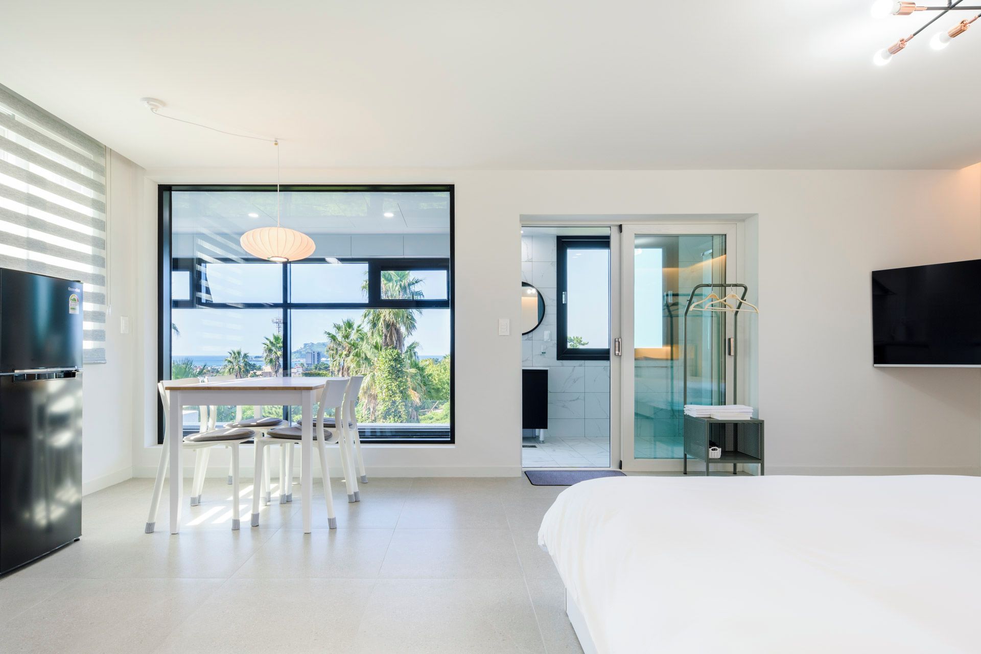 Un dormitorio moderno con una cama grande, una mesa de comedor con sillas, un televisor montado en la pared y vistas a palmeras a través del ventanal.