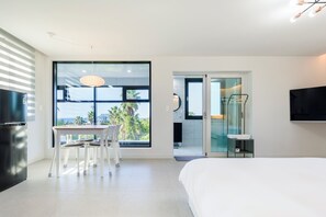A modern bedroom with a large bed, a dining table with chairs, a TV mounted on the wall, and a view of palm trees through the window. - Liverpool an spa (Jeju City)