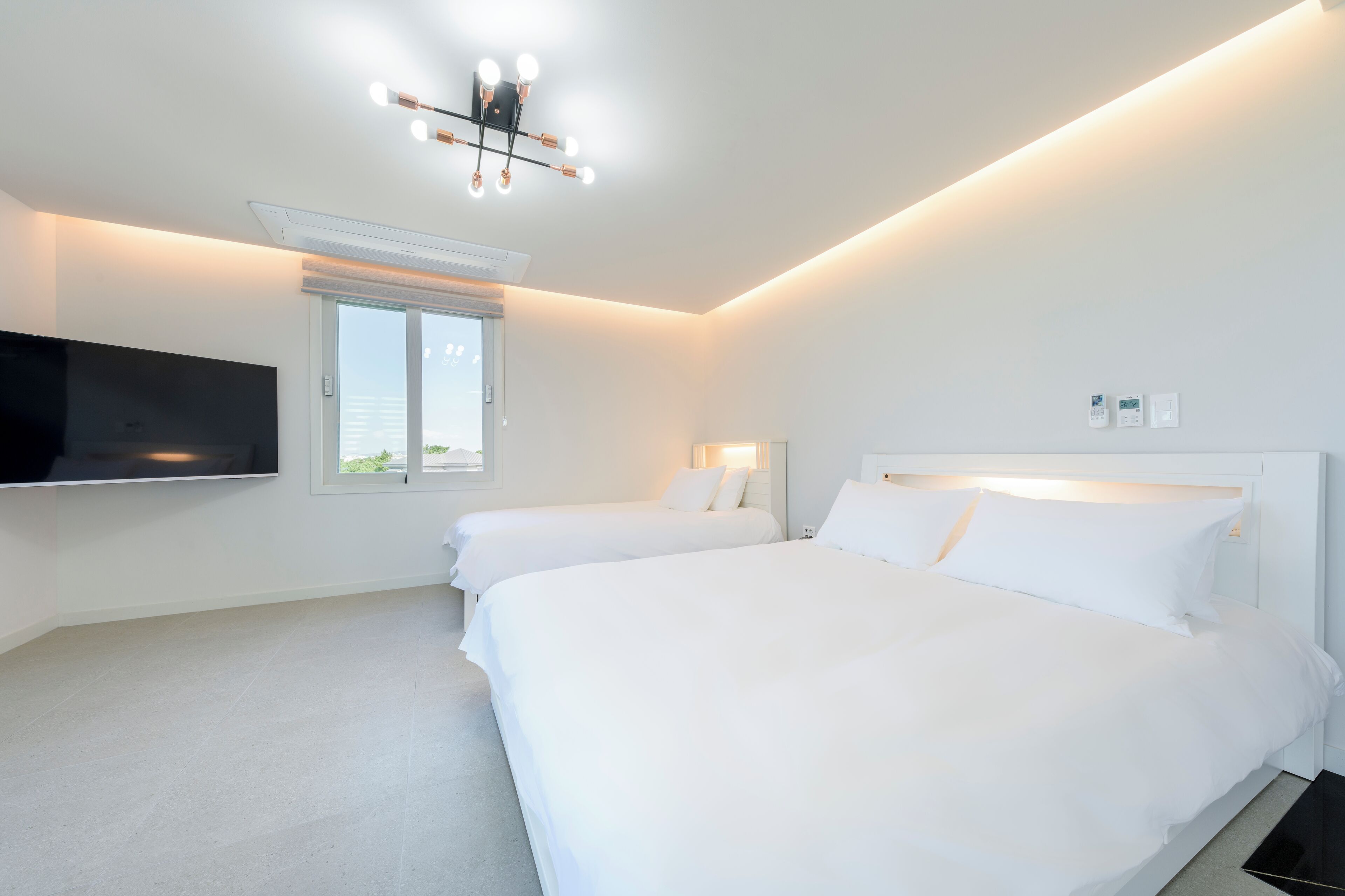A hotel room with two beds, a flat-screen TV mounted on the wall, a window, and a ceiling light fixture.