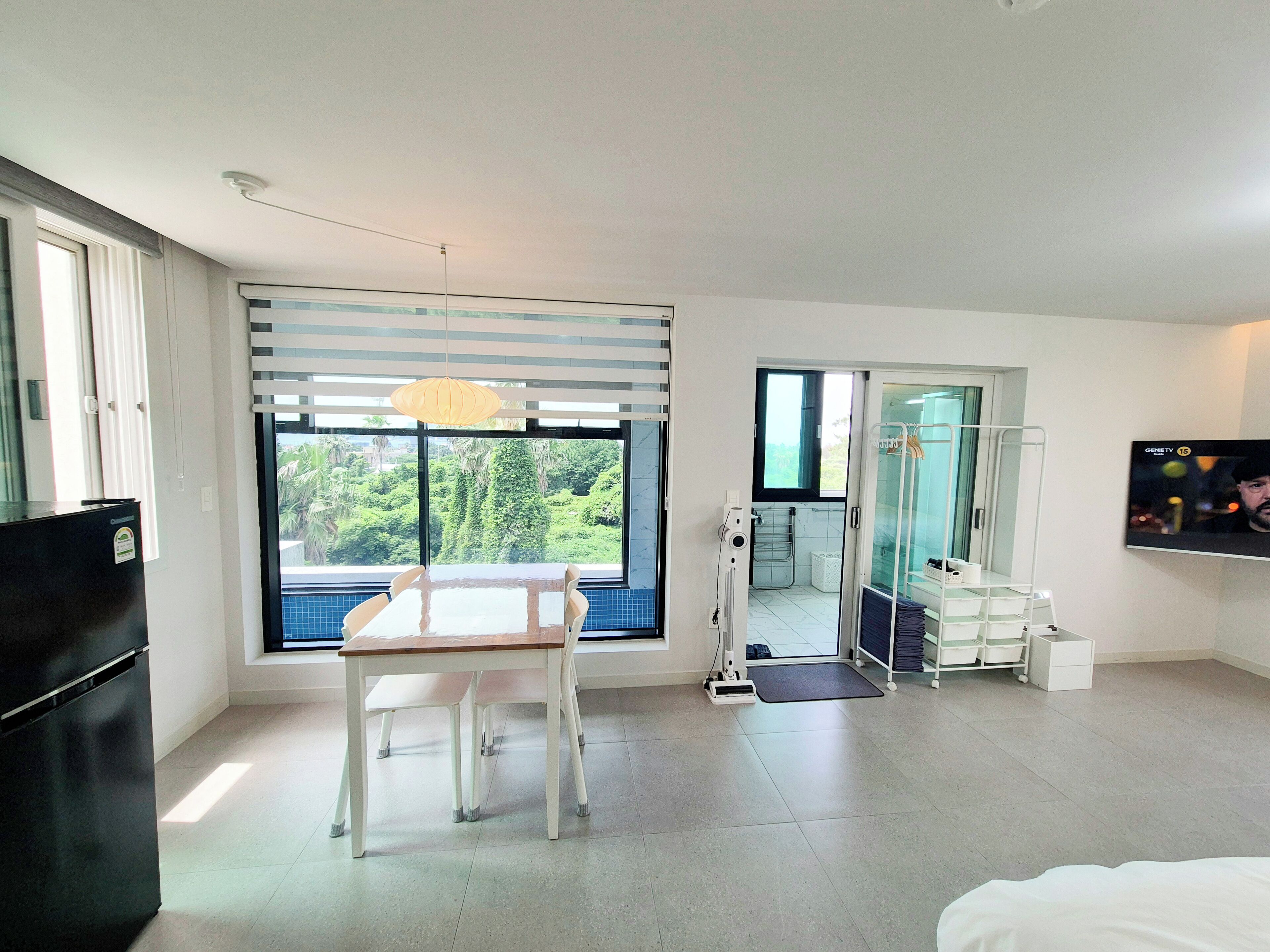 Una habitación moderna con un gran ventanal, una mesa de comedor, un refrigerador negro, cabina de ducha de vidrio y un televisor de pared.