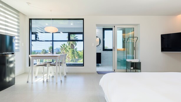 A modern hotel room with a large window, a bed, a TV, and a bathroom with a glass door. - Liverpool an spa (Jeju City)