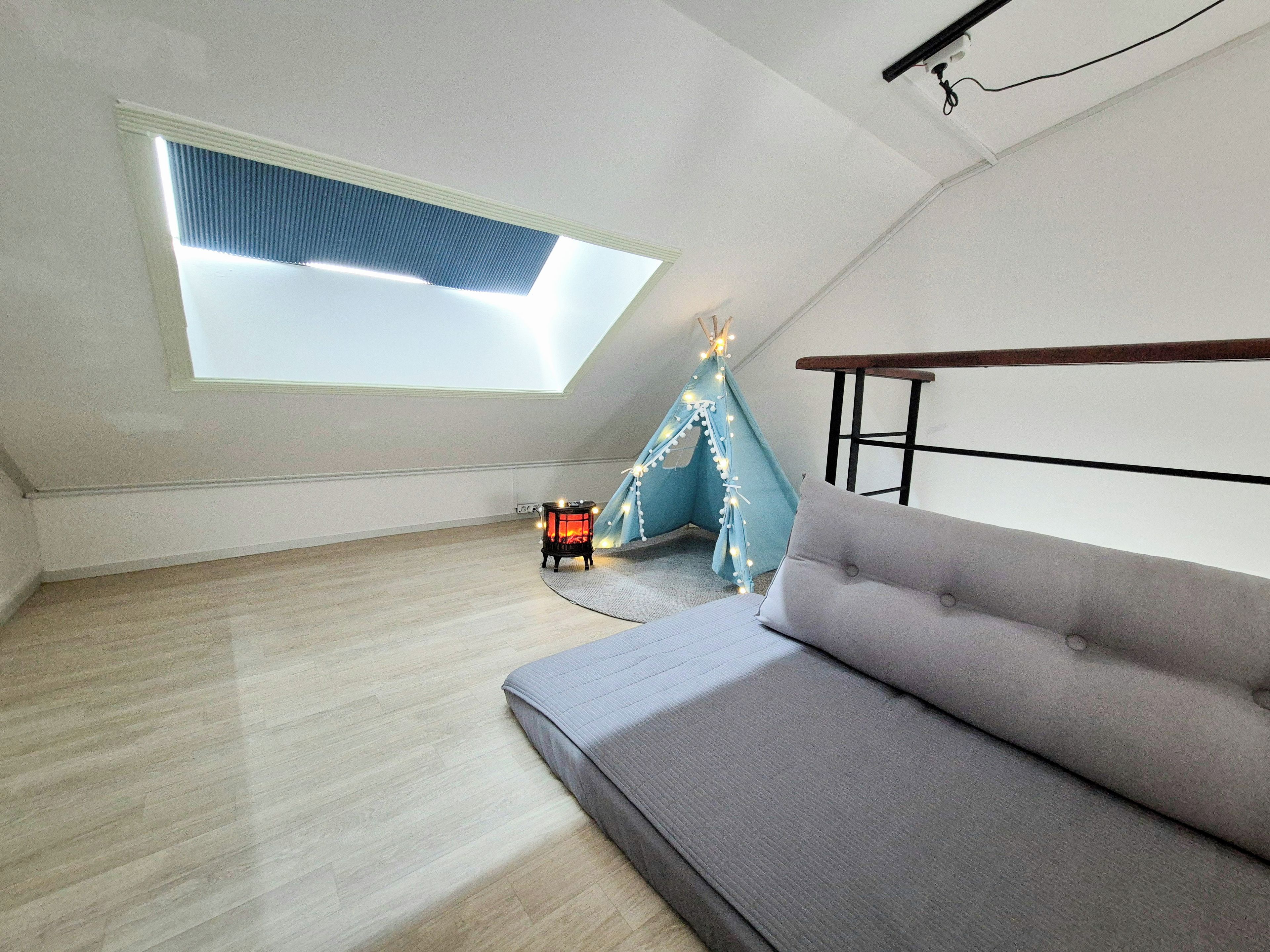 A loft bedroom with a skylight, a small heater, a blue teepee, and a grey sofa.