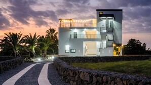 A modern two-story house with a stone retaining wall, a paved driveway, and a string of lights. - Liverpool an spa (Jeju City)