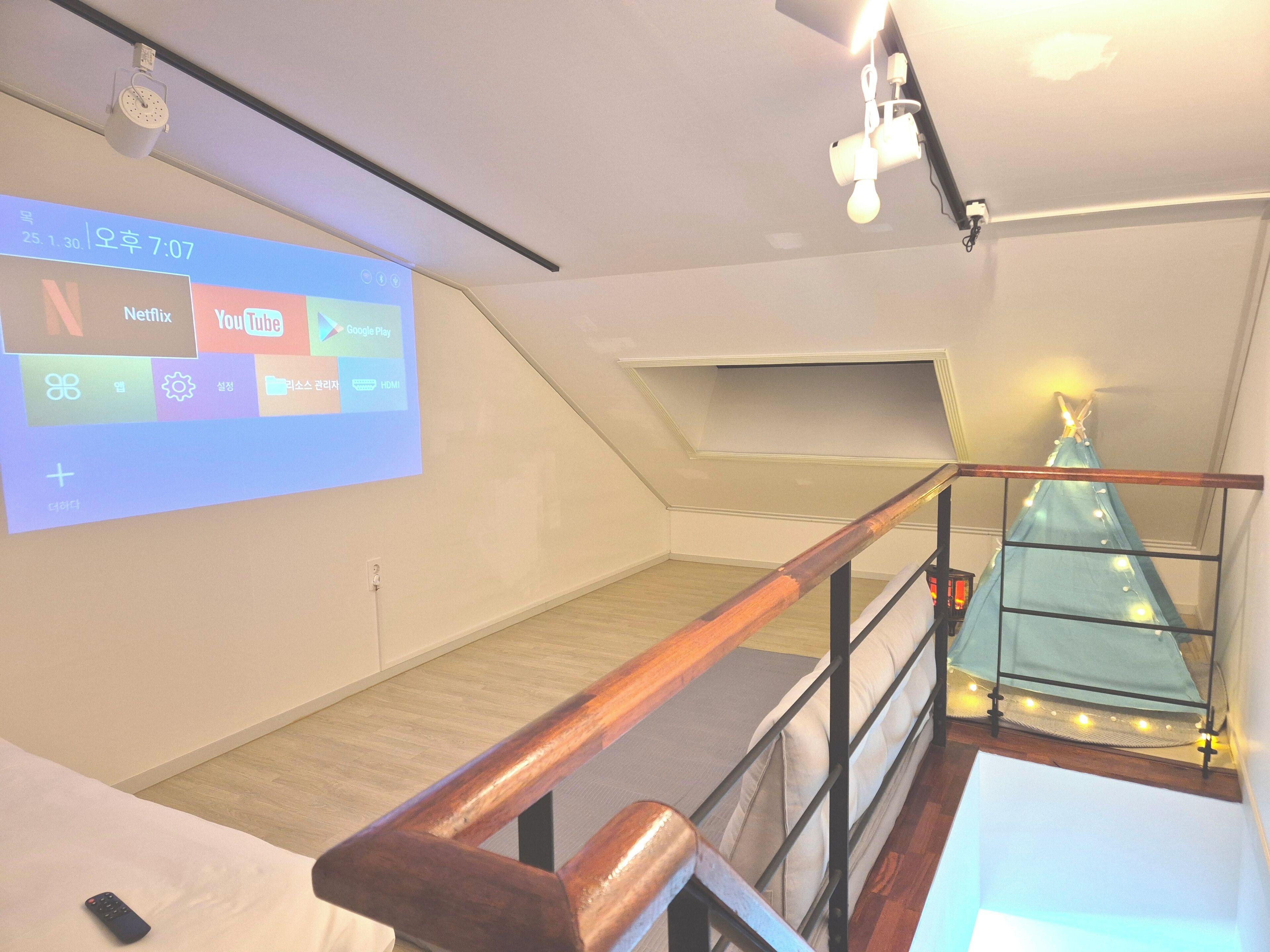 Ein modernes Schlafzimmer im Loft-Stil mit schrägem Dach, einer Holztreppe, einer Projektionsleinwand, die Streaming-Dienste zeigt, und einem Weihnachtsbaum.