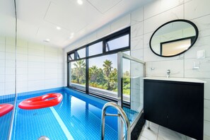 An indoor swimming pool with clear blue tiles, a black and white tiled wall, a large window with a view of palm trees, and a black cabinet with a round mirror. - Liverpool an spa (Jeju City)