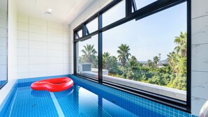 An indoor swimming pool with blue tiles, a red inflatable ring, and a view of palm trees and buildings through large windows. - Liverpool an spa (Jeju City)