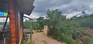 A rustic wooden building with a dirt path leading to a gate, surrounded by dense greenery and trees.