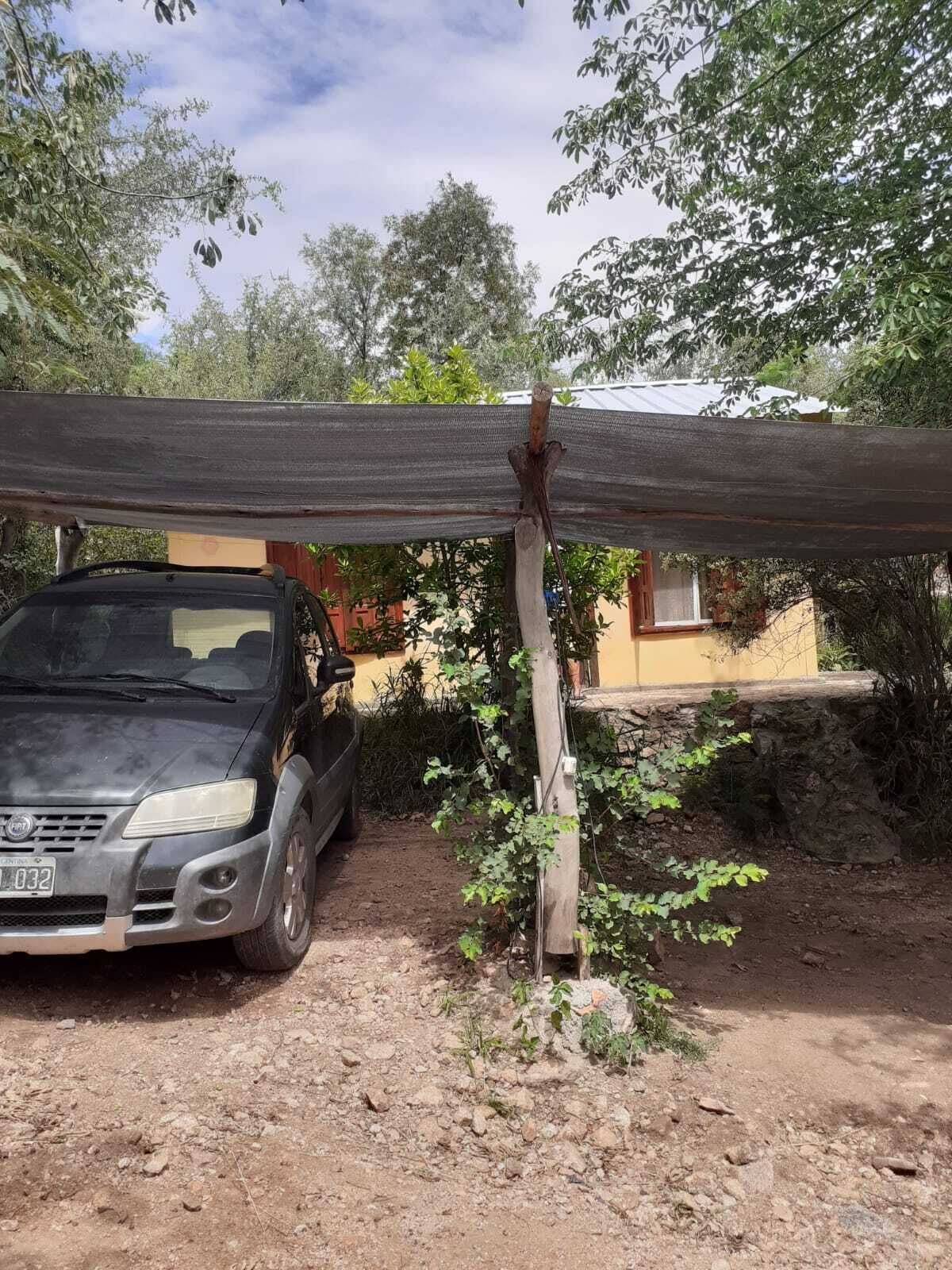 Un vehículo estacionado bajo una estructura improvisada, un terreno de tierra y un edificio con fachada amarilla al fondo.