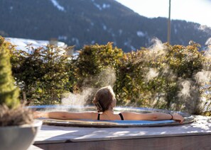 A person relaxing in a hot tub with steam rising, surrounded by greenery and mountains in the background. - No.14 Verbier (Bagnes)