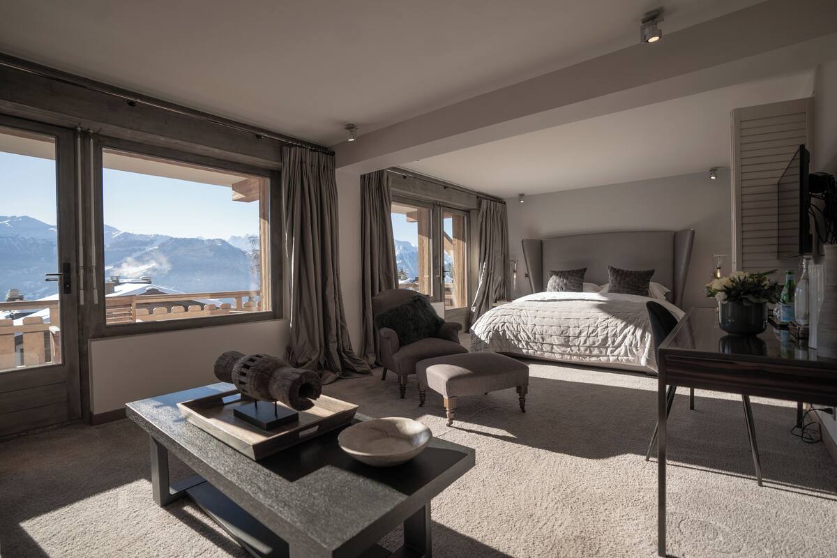 A spacious bedroom with a large bed, a desk, and a view of mountains through large windows.