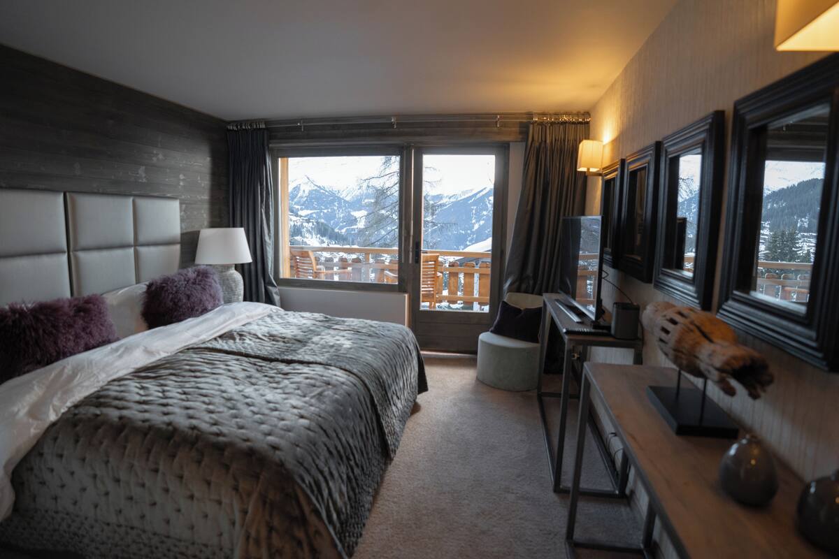 A bedroom with a large bed, a desk, a chair, a TV, and a view of snow-capped mountains.