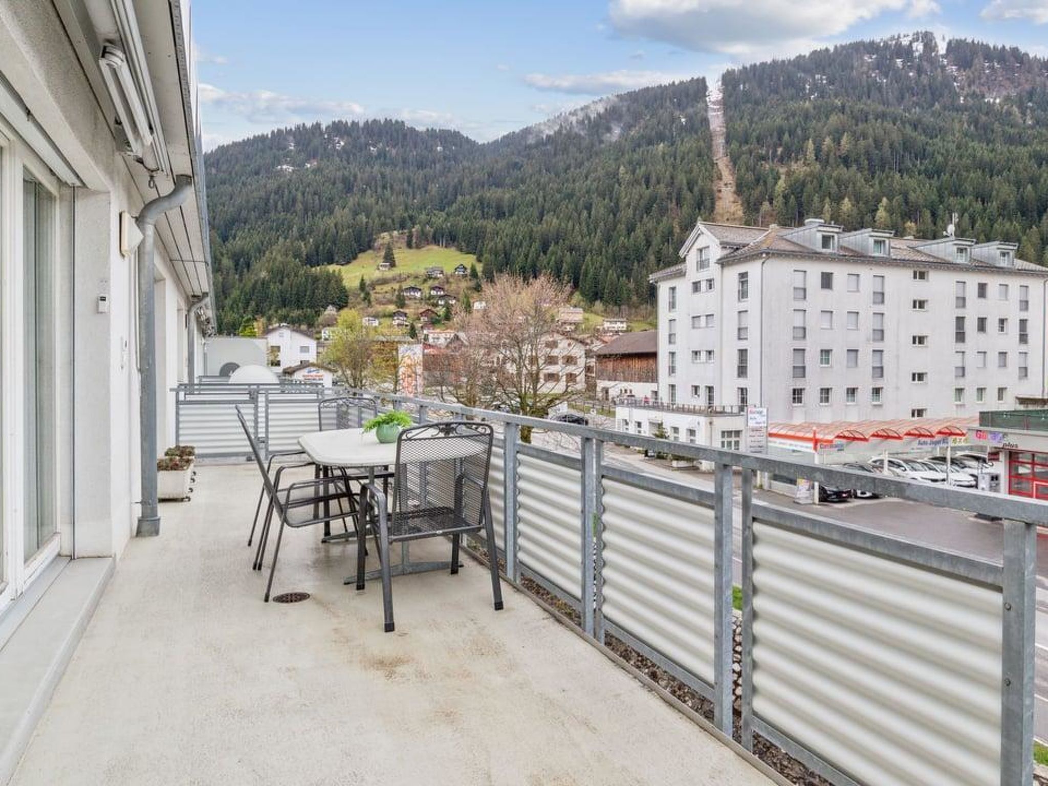 En balkon med et bord og stole, med udsigt over et bjerglandskab og en by med bygninger.