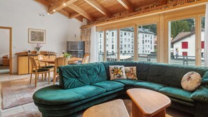 A cozy living area with a green sectional sofa, wooden coffee table, and a dining table with chairs. A television and a fireplace are visible in the background. - Casa Hermi by Interhome (Churwalden)