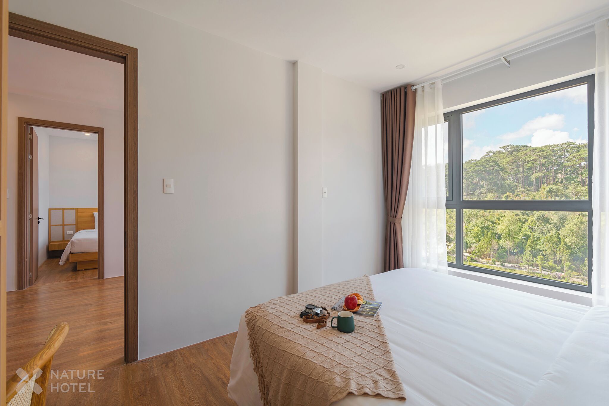 Habitación de hotel con una ventana grande, una cama, una mesita de noche con una cámara y una taza, y vistas a un bosque.