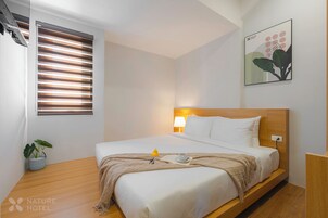 Habitación de hotel con una cama, una lámpara de noche, una ventana con persianas, una obra de arte en la pared y una pequeña planta en maceta.