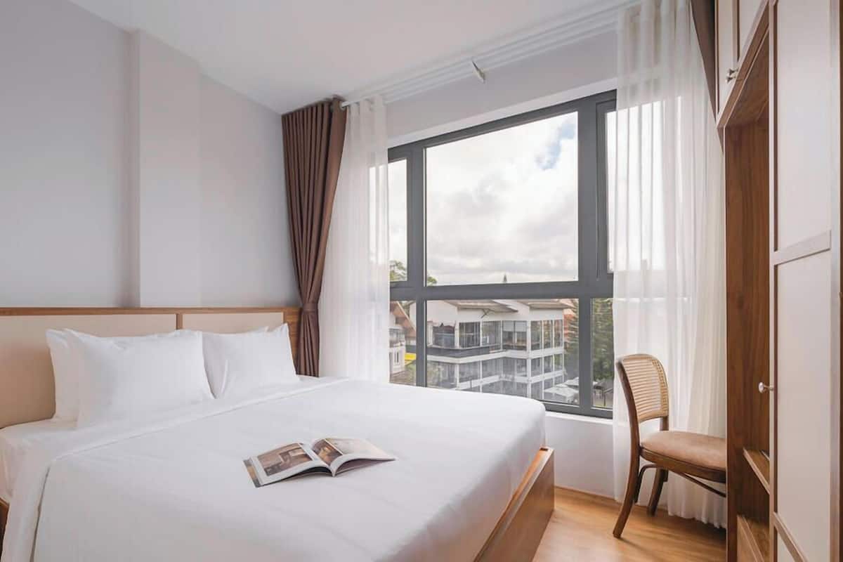 A hotel room with a bed, a window with curtains, a chair, and a nightstand with a lamp.