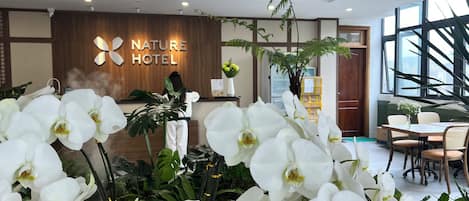 A hotel reception area with a 'Nature Hotel' sign, a receptionist, and a reception desk. There are white orchids and a large potted plant in the foreground.