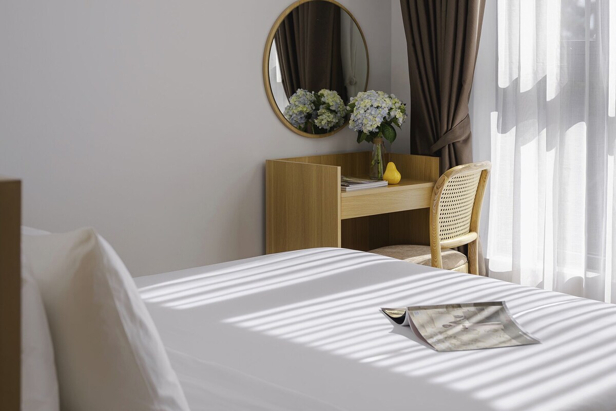 A hotel room with a bed, a desk with a chair, a mirror, and a window with curtains.