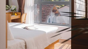 Une chambre avec une grande fenêtre, un lit, une table de chevet avec un miroir et un petit bureau avec une chaise.