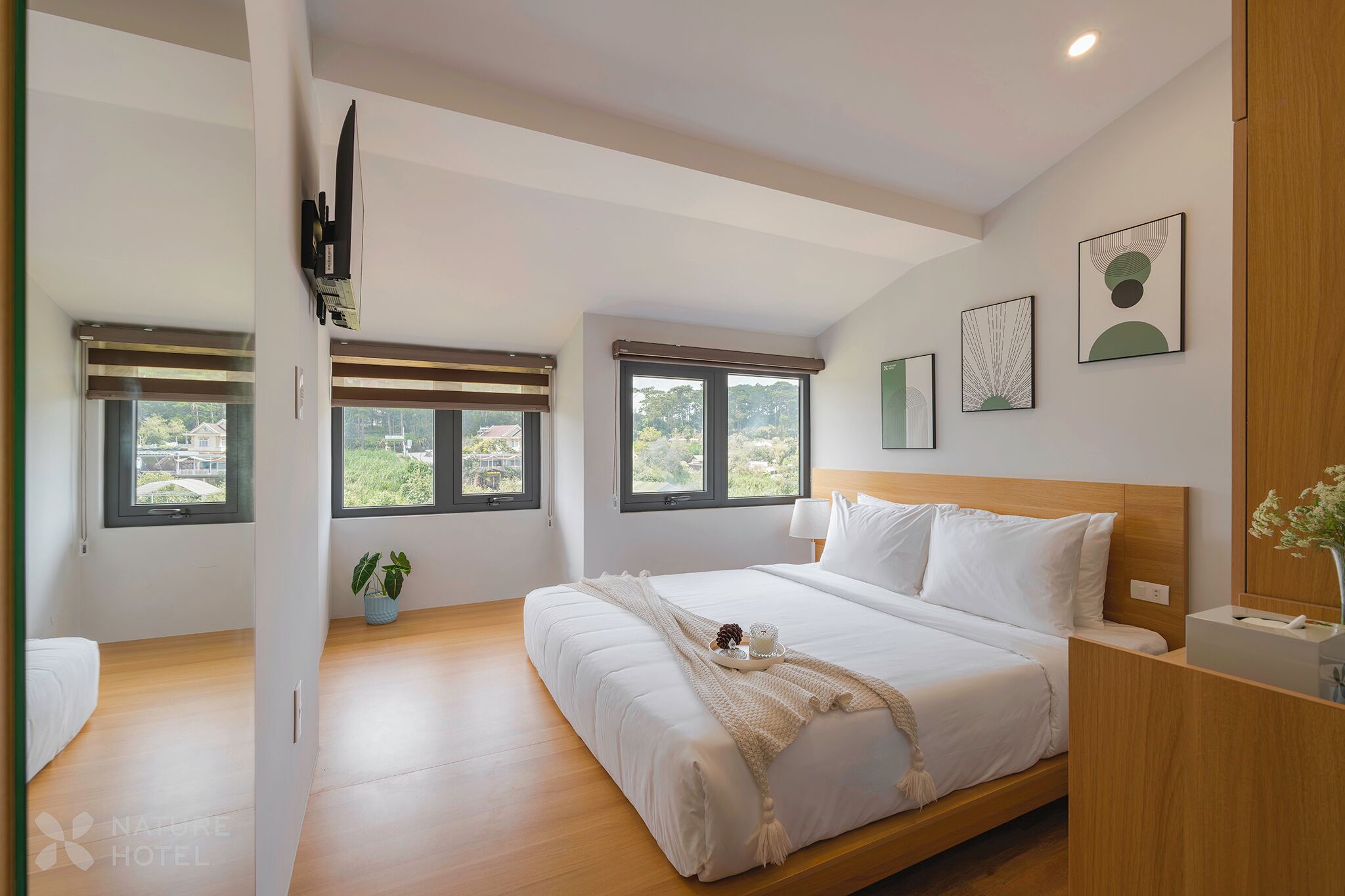 A modern hotel room with a large bed, two windows, and a view of greenery.