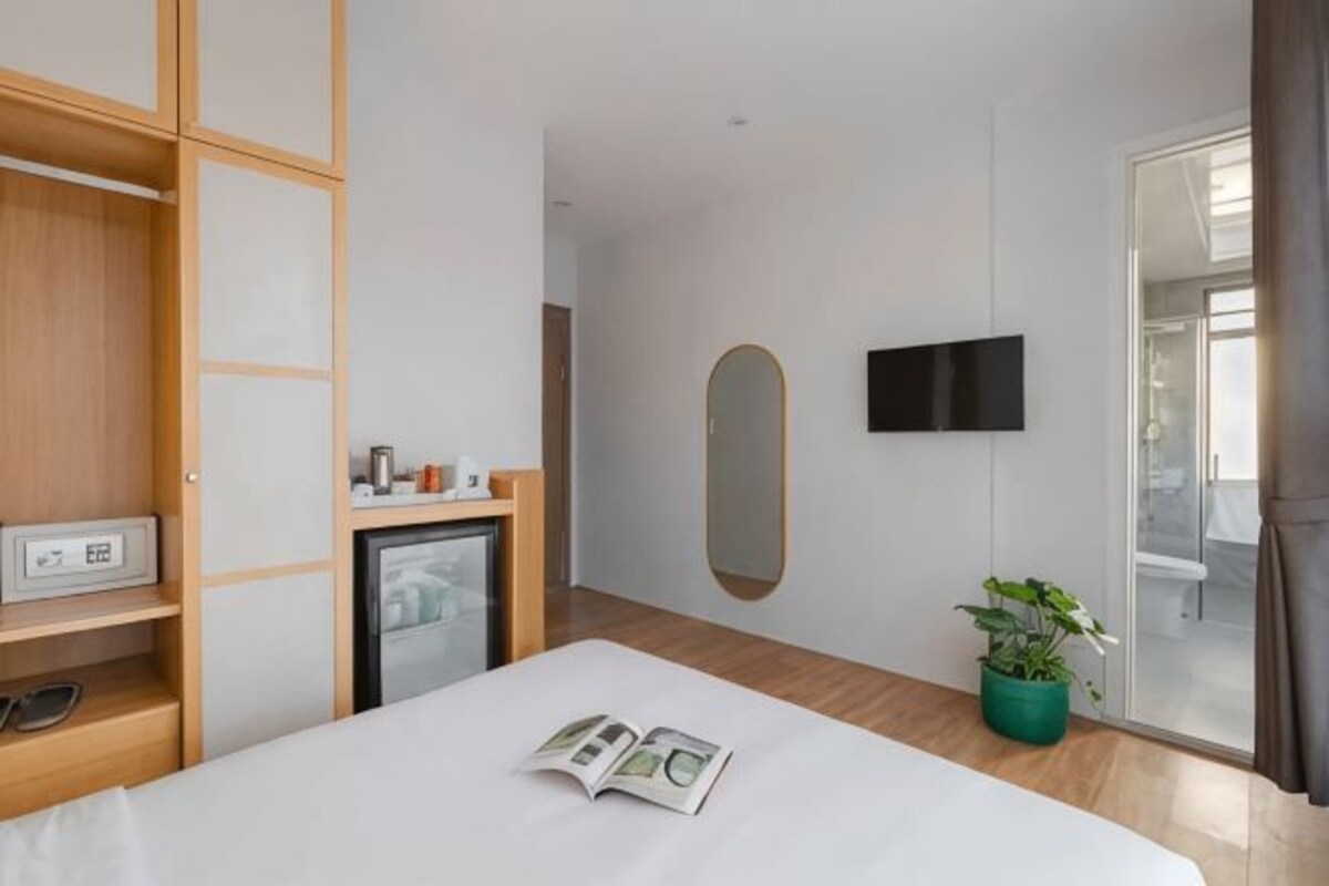 A hotel room with a bed, a television mounted on the wall, a small refrigerator, a wooden wardrobe, and a potted plant.
