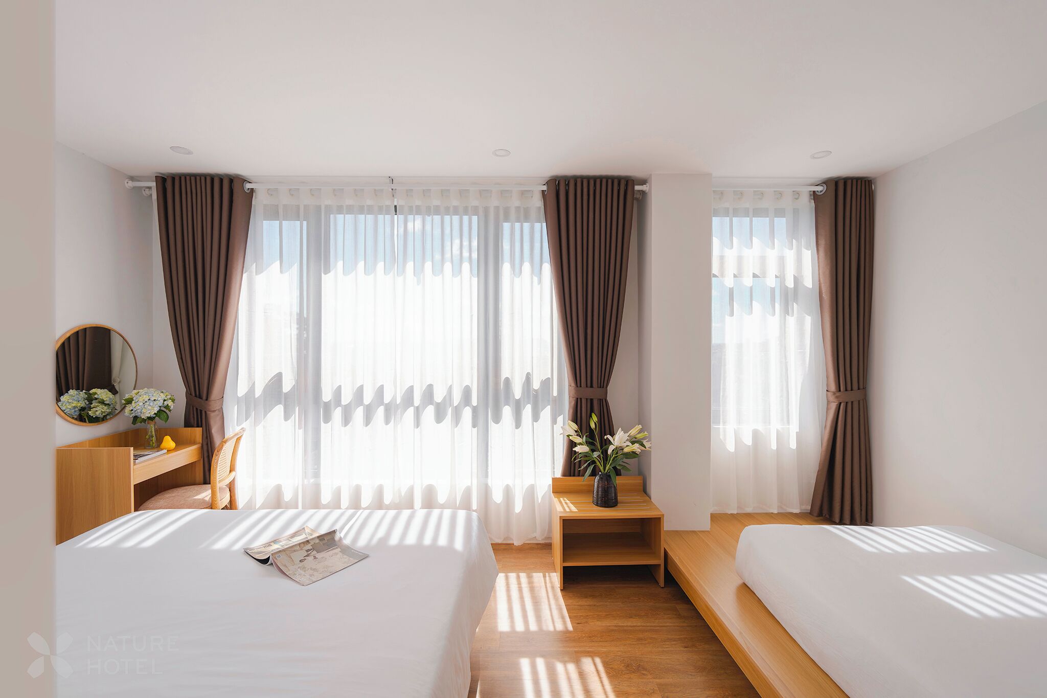 A hotel room with two beds, a wooden nightstand, a small table with a vase of flowers, and a mirror on the wall.