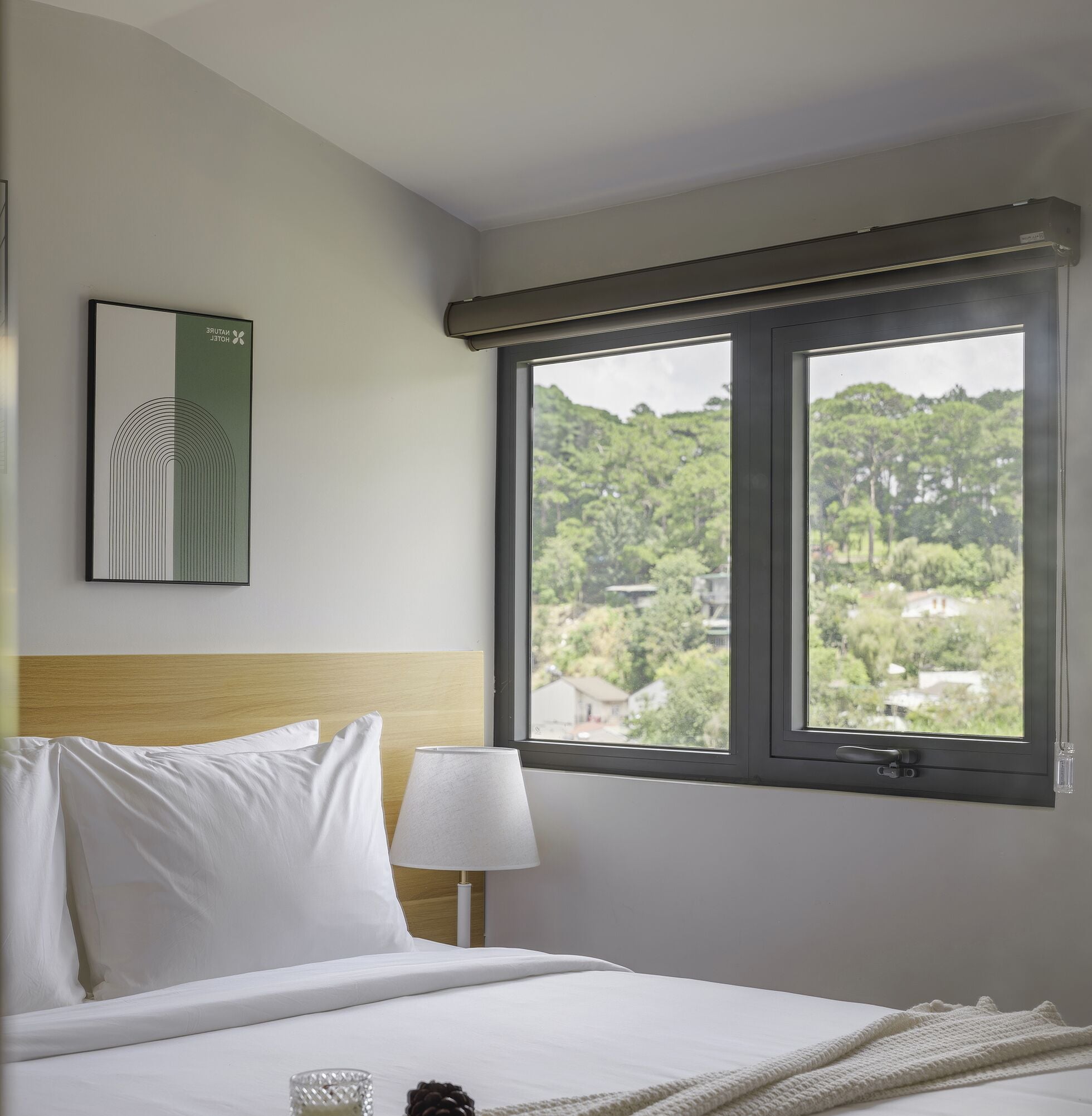 Una habitación de hotel con una cama, una mesita de noche, una lámpara, un cuadro enmarcado y una ventana con vistas a zonas verdes y edificios.