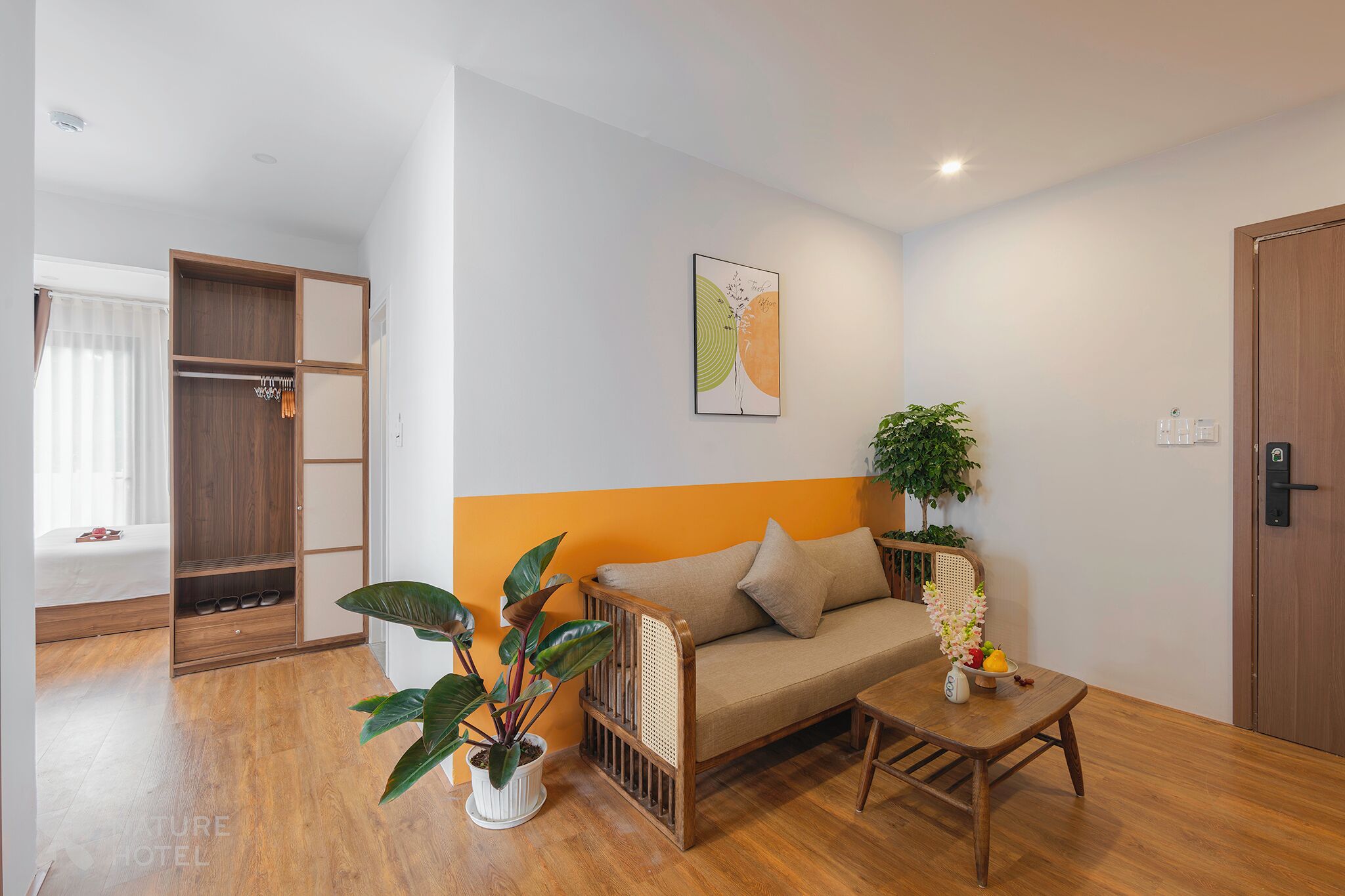 A room with a wooden floor, a sofa with cushions, a small table with a vase, a potted plant, a wardrobe, and a bed with a curtain.