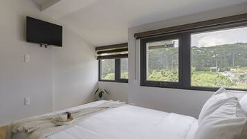 A bedroom with a bed, a TV mounted on the wall, a window with a view of greenery, and a bedside table with a lamp.