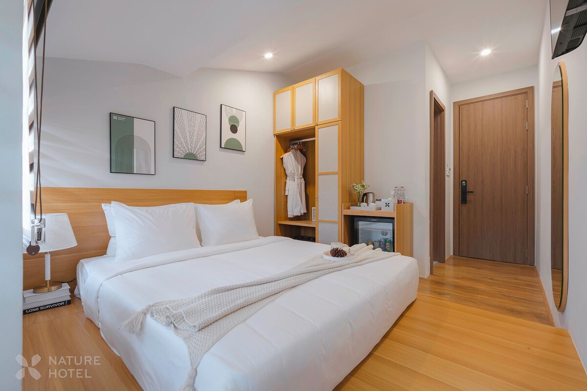 A hotel room with a large bed, wooden headboard, a wardrobe, a desk with a chair, and a mirror.