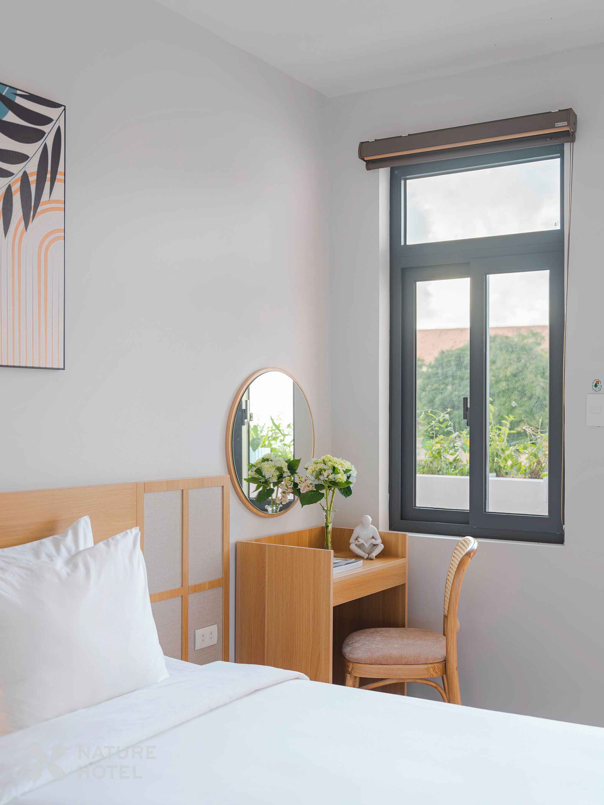 A hotel room with a bed, a wooden desk, a chair, a mirror, and a window with a view of greenery.