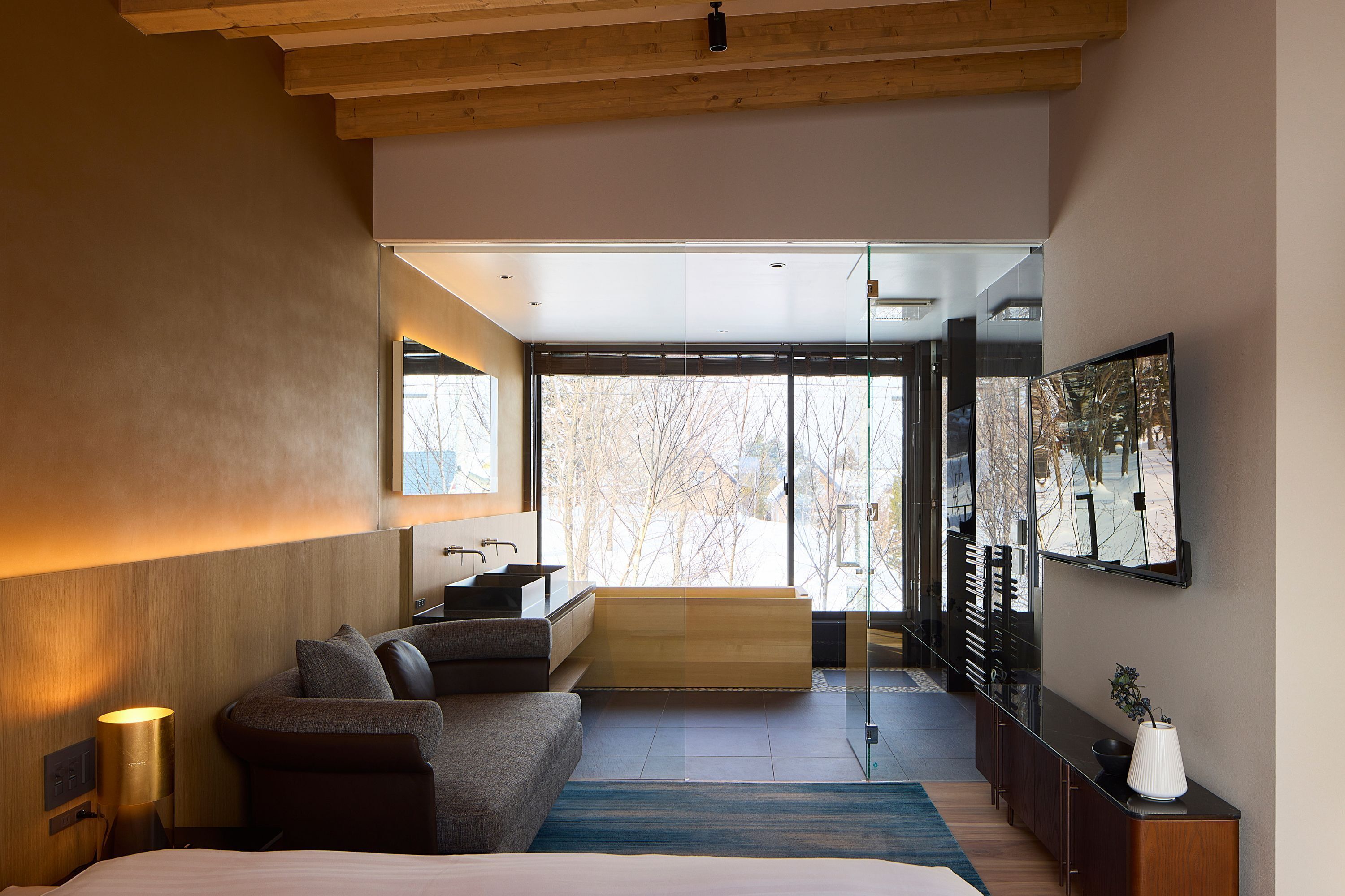 A modern hotel room with a large bed, a sofa, a small kitchen area, and a large window offering a view of trees.