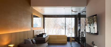 A modern hotel room with a large bed, a sofa, a small kitchen area, and a large window offering a view of trees.