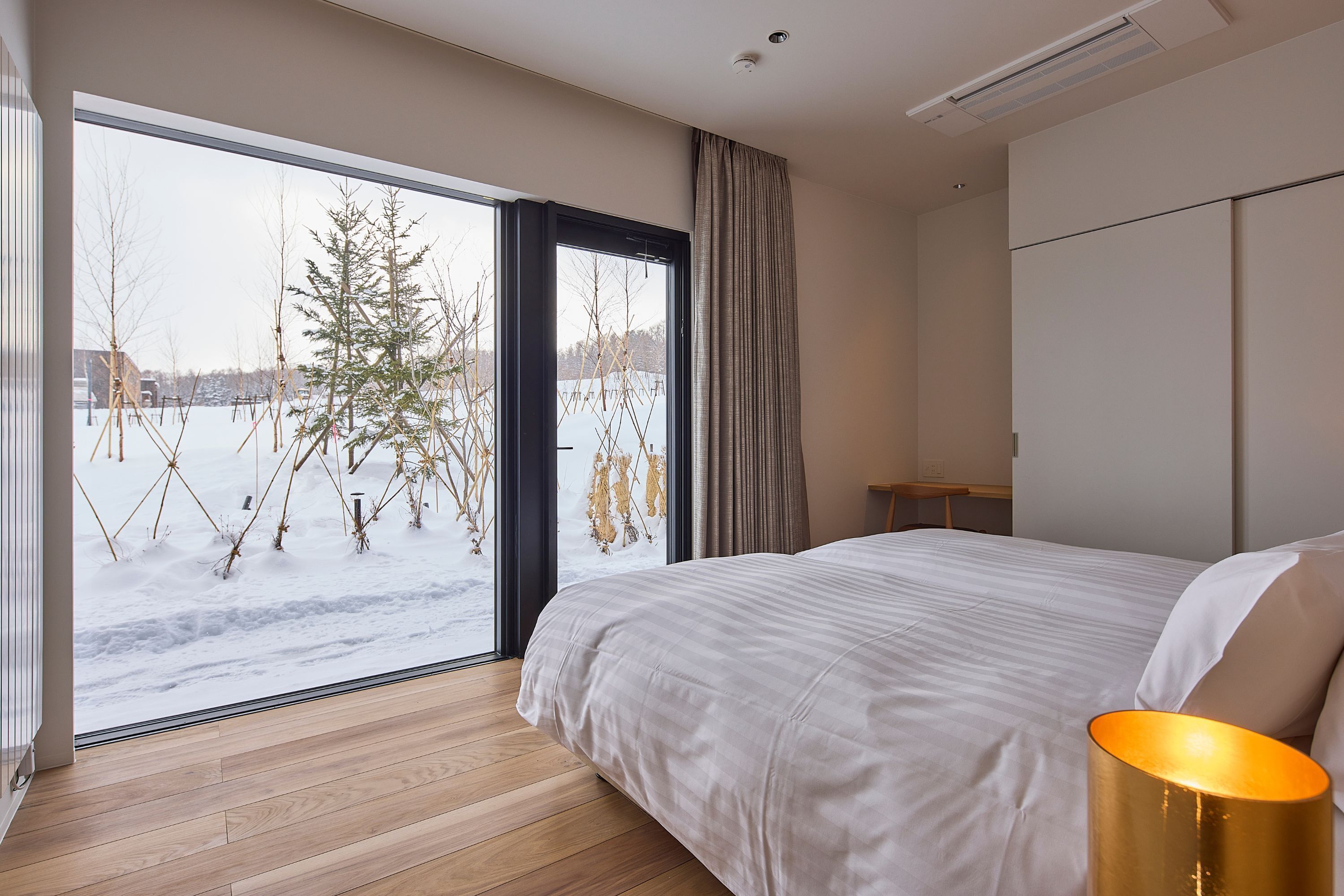 A bedroom with a large window, a bed, a bedside table, and a lamp.