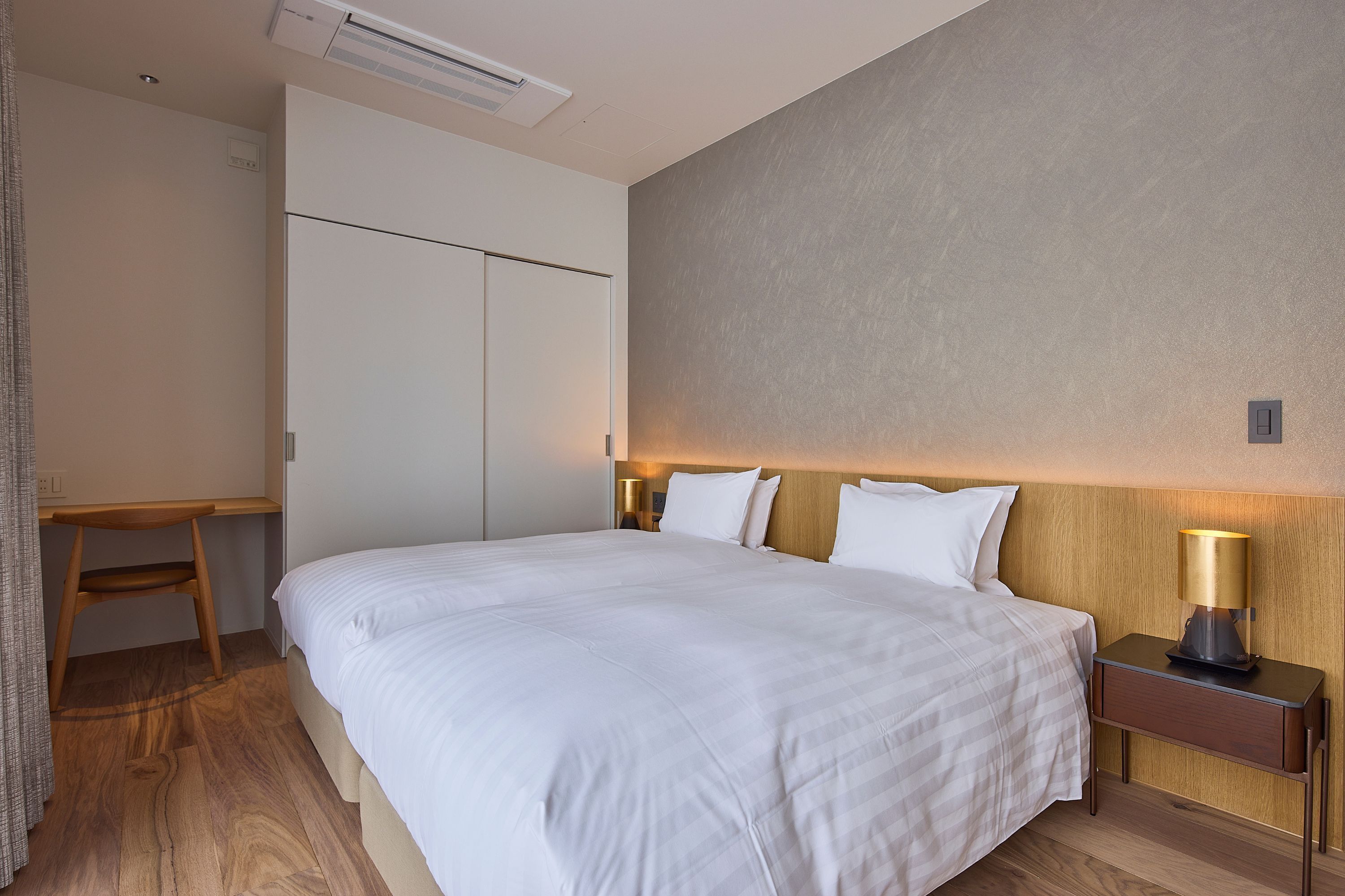 Una habitación de hotel con una cama grande, un escritorio de madera, una mesita de noche con lámpara y un armario.