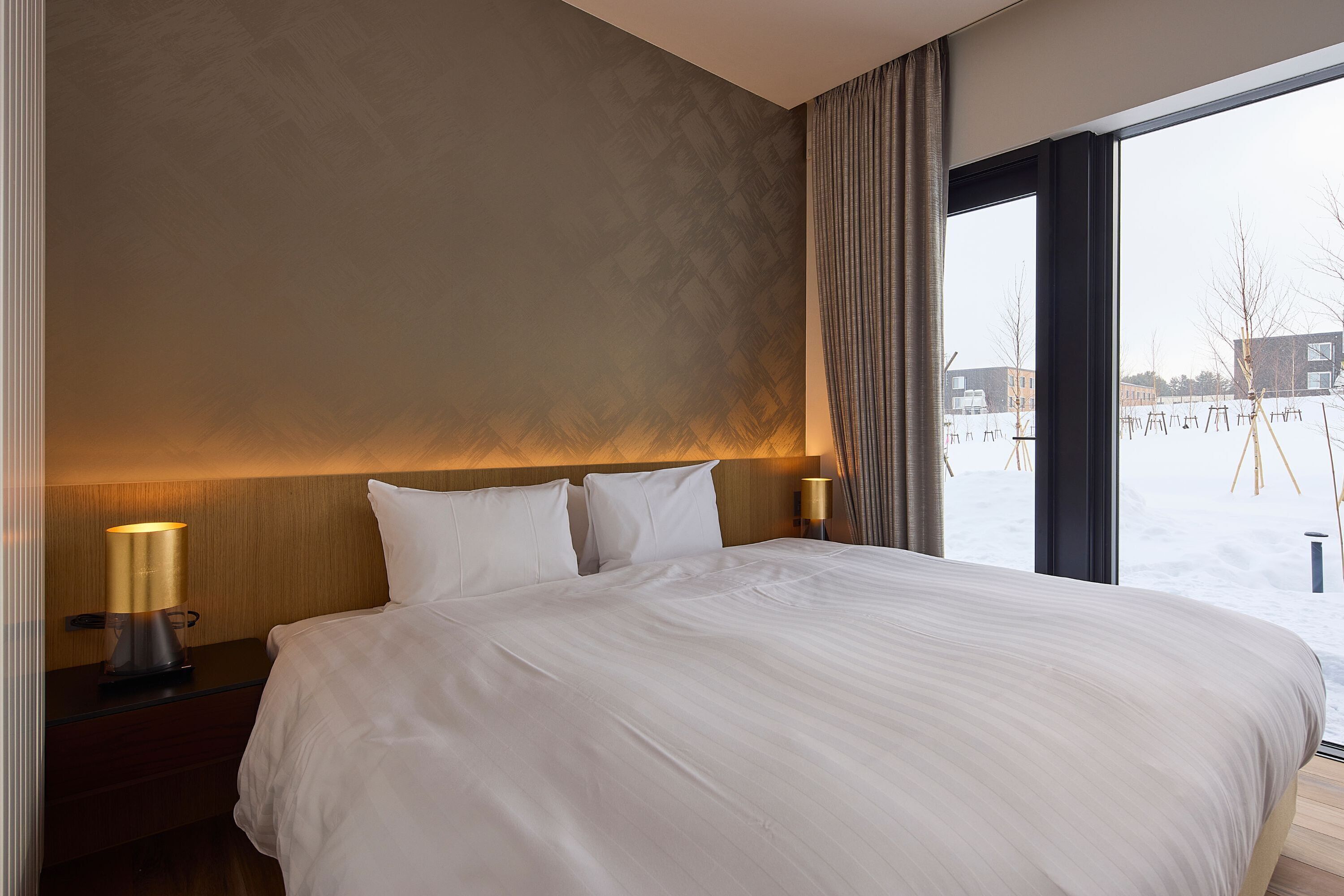 Une chambre d’hôtel avec un grand lit, une table de chevet avec une lampe et une vue sur un paysage enneigé par une grande fenêtre.