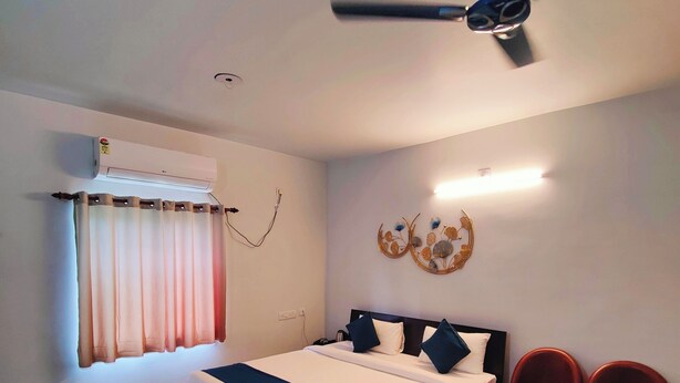 A hotel room with a bed, a desk, two chairs, a ceiling fan, an air conditioning unit, and a decorative wall piece. - The Nook (Bhubaneshwar)