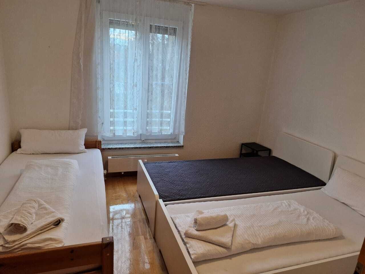 A small, clean room with two beds, a window with sheer curtains, and a radiator.