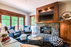 A cozy living room with a stone fireplace, comfortable seating, and large windows offering a view of greenery.