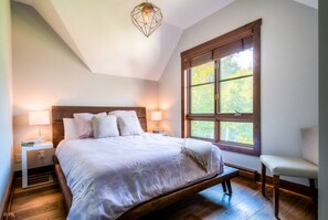 A bedroom with a large bed, two bedside lamps, a window with a view of trees, and a wooden floor.