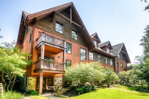 A large, multi-story wooden house with a balcony on the first floor, surrounded by lush greenery.