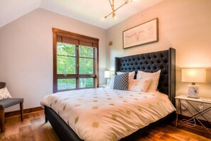 A bedroom with a large bed, a window, a chair, and a bedside table with a lamp.