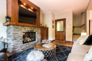 A cozy living room with a stone fireplace, a flat-screen TV mounted above it, and a comfortable seating area with a round coffee table.