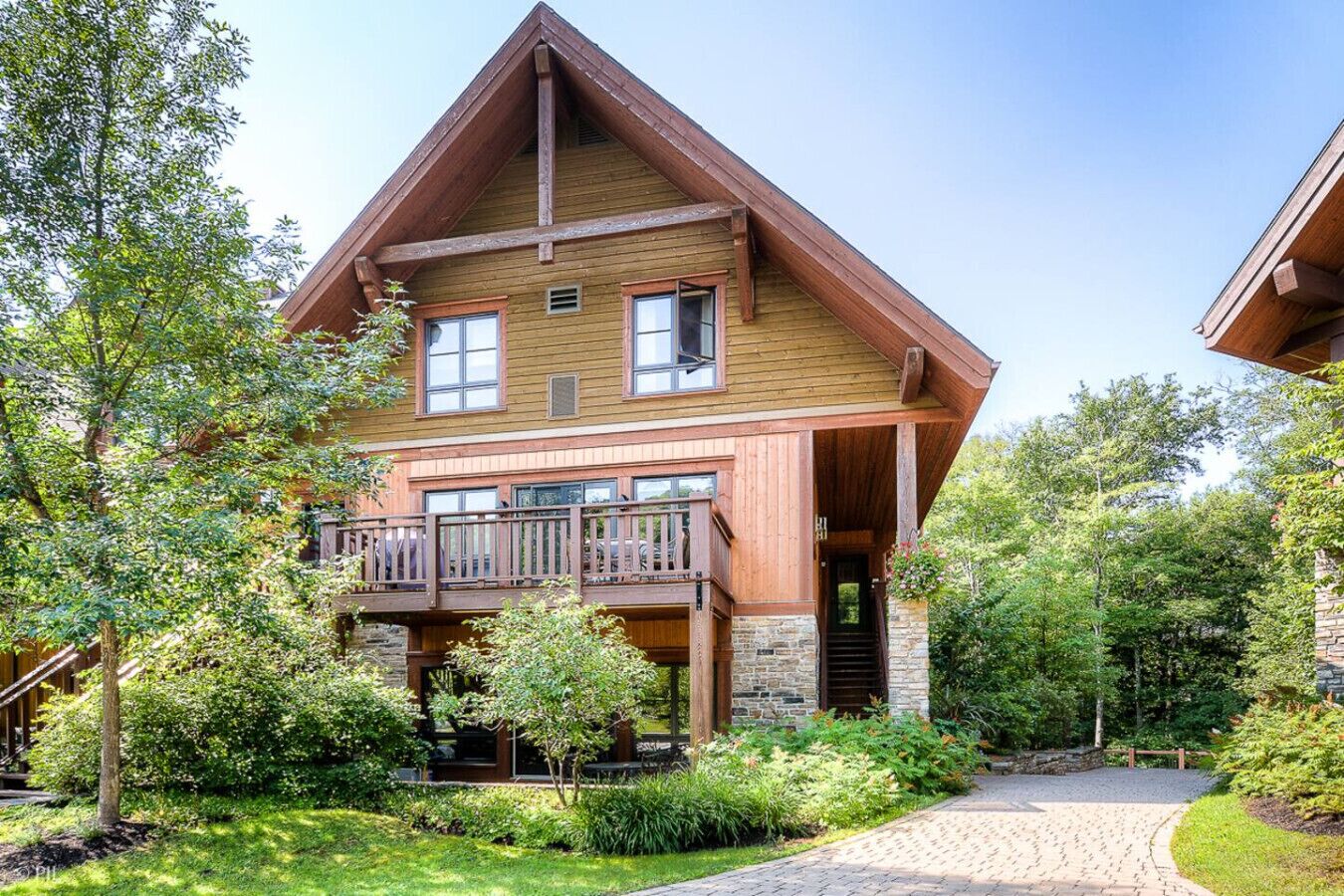 A two-story wooden house with a balcony, surrounded by lush greenery and a stone pathway.