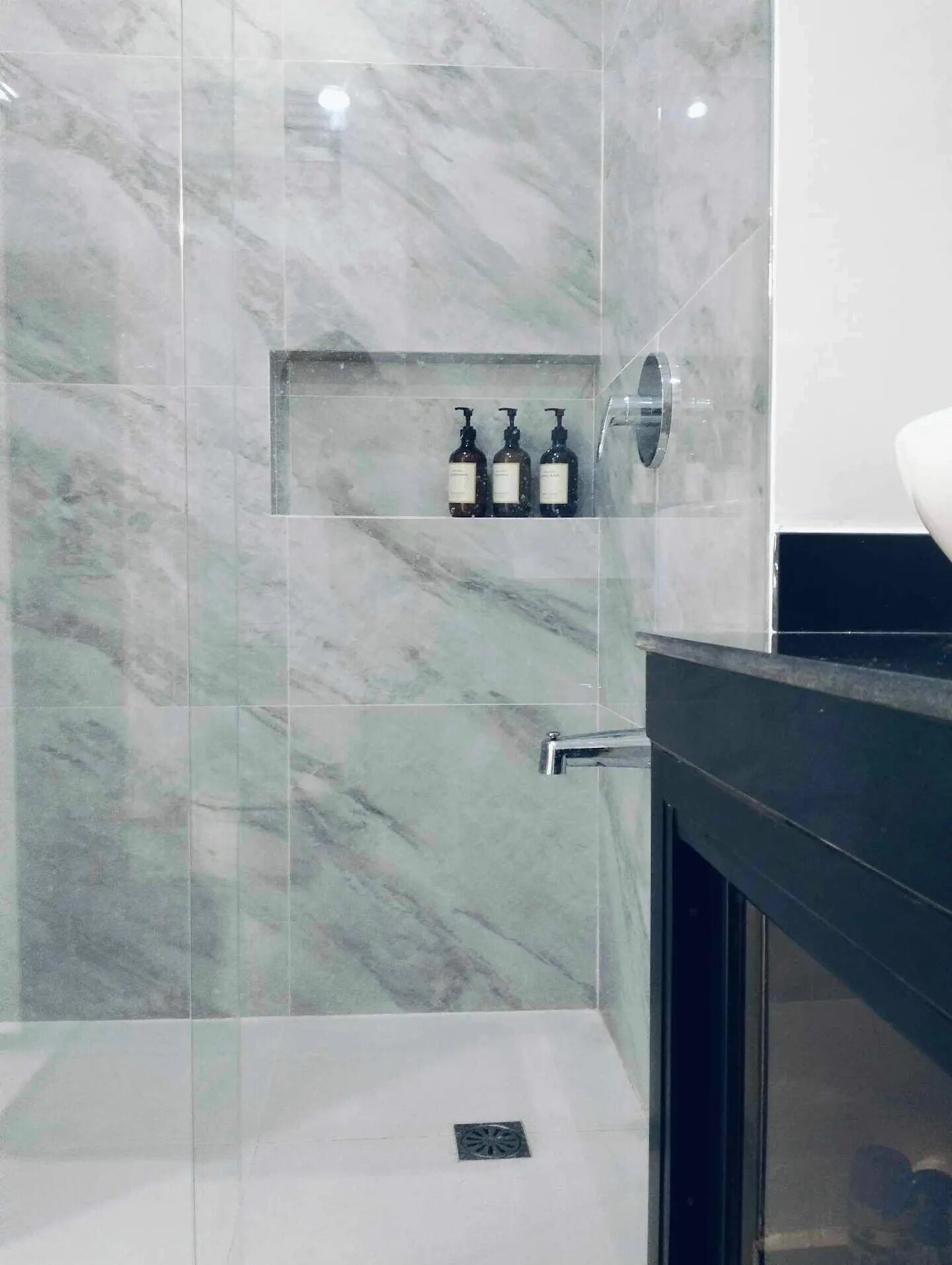 A bathroom with marble walls, a glass shower enclosure, and three bottles of shampoo on a shelf.