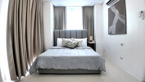 A modern bedroom with a large bed, grey curtains, a bedside table with a lamp, and a wall-mounted air conditioning unit. - Stay in Unit 301 Pontefino Tower, a Cozy 1 Bedroom Apartment that is Brand New (Batangas)